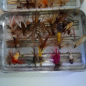 May include: A silver metal fly fishing box containing an assortment of artificial fishing flies. The flies feature various colors including red, yellow, brown, white, and black, with intricate feather and hair details attached to small metal hooks.
