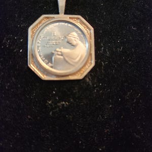 May include: Silver pendant with an octagonal frame and a round image of a person in a robe holding a book. The image is surrounded by a circle of text.
