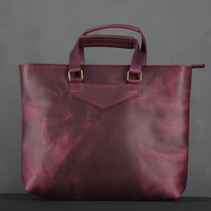 May include: A burgundy leather tote bag with a rectangular shape and two handles. The bag has a small pocket on the front and gold-colored metal hardware. The bag is made of a smooth, textured leather.
