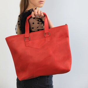 May include: A bright red leather tote bag with two handles and brass-colored hardware. The bag has a rectangular shape with a structured design. The bag is being held by a person.