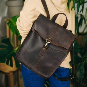 May include: A brown leather backpack with a flap closure and a metal buckle. The backpack has a front pocket and adjustable straps.
