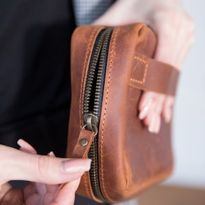 Peut inclure: Une pochette en cuir marron zippée avec une fermeture éclair noire et une sangle en cuir marron. La pochette est tenue par une main.