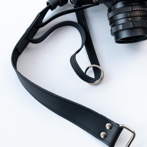 Peut inclure: Sangle de caméra en cuir noir avec un anneau en métal argenté et une boucle en métal argenté. La sangle est attachée à un appareil photo noir.