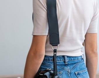 Peut inclure: Une personne portant une chemise blanche et un jean bleu porte une bandoulière d'appareil photo noire avec un appareil photo Canon EOS Rebel.