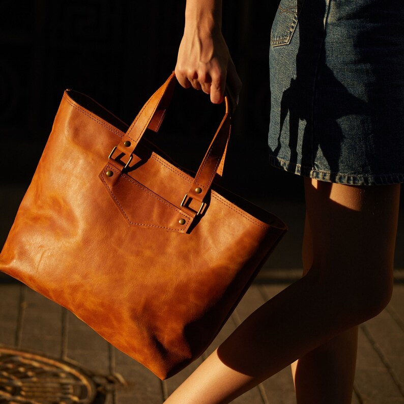 May include: A large, rectangular, tan leather tote bag with two top handles and a front pocket. The bag features brass-colored hardware and is held by a person. The bag is made of a smooth, durable leather.