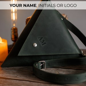May include: A dark green leather triangular crossbody bag with a silver buckle and a silver button. The bag has the initials "AM" embossed on the front.