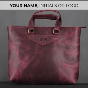May include: Burgundy leather tote bag with top handles and a shoulder strap. The bag features gold-colored hardware and the initials "MN" embossed on the front. The text "YOUR NAME, INITIALS OR LOGO" is at the top.