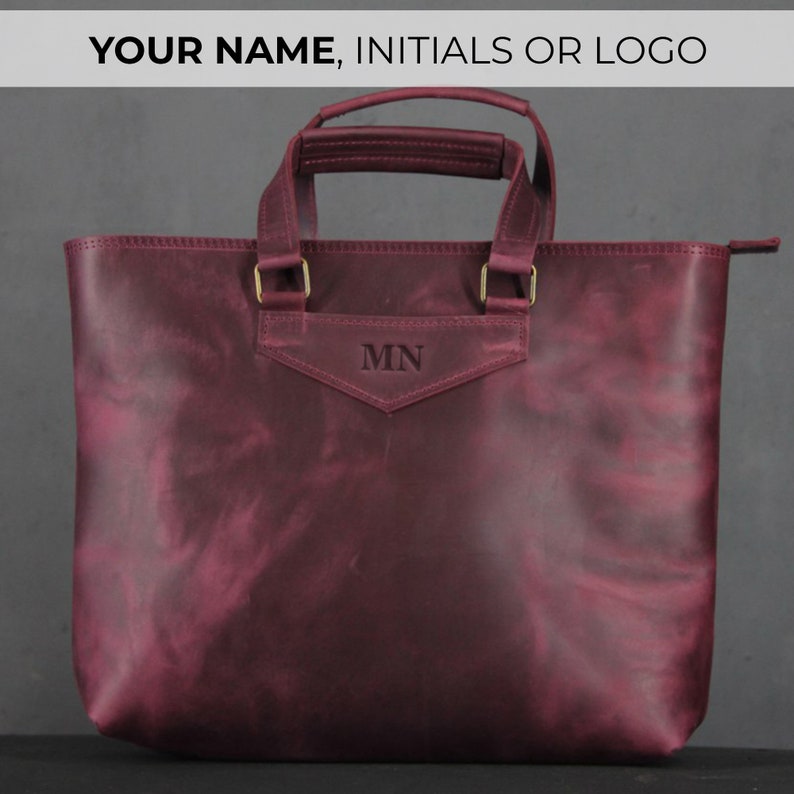 May include: Burgundy leather tote bag with top handles and a shoulder strap. The bag features gold-colored hardware and the initials "MN" embossed on the front. The text "YOUR NAME, INITIALS OR LOGO" is at the top.