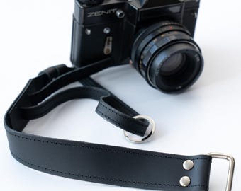 Peut inclure: Une sangle de caméra en cuir noir avec un anneau et une boucle en métal argenté. La sangle est attachée à un appareil photo noir avec le mot "ZENIT" imprimé dessus.
