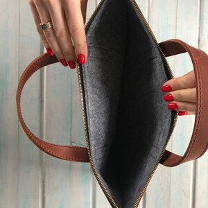 Puede incluir: Un bolso de cuero marr&oacute;n para port&aacute;til con cierre de cremallera y forro de fieltro gris. El bolso tiene dos asas superiores y un bolsillo con cremallera en la parte delantera.