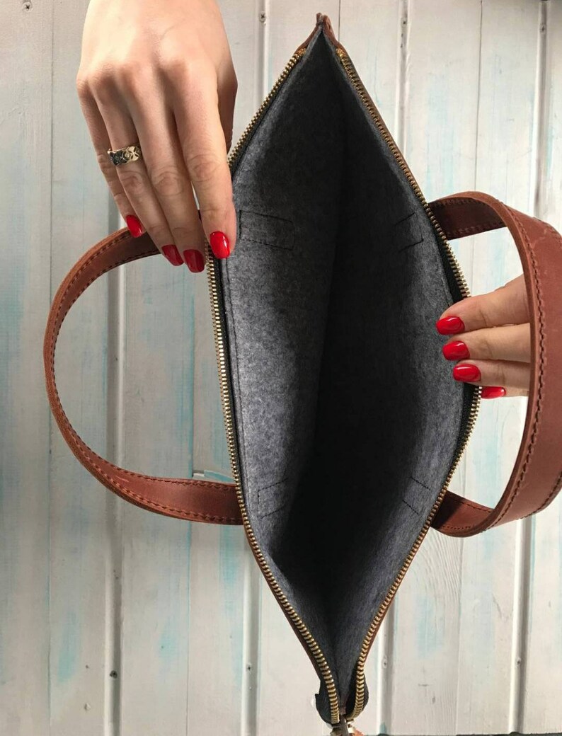 Puede incluir: Un bolso de cuero marr&oacute;n para port&aacute;til con cierre de cremallera y forro de fieltro gris. El bolso tiene dos asas superiores y un bolsillo con cremallera en la parte delantera.
