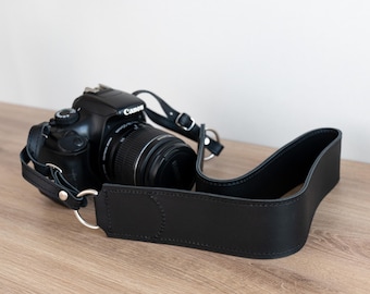 Peut inclure: Un appareil photo noir avec une sangle en cuir noir. L'appareil photo a un objectif avec une bague argentée et le mot "Canon" imprimé sur le dessus.
