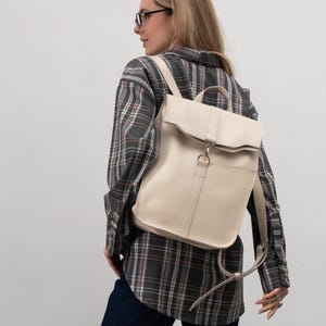 May include: A cream-colored leather backpack with a flap closure and a silver buckle. The backpack has a single pocket on the front and is worn by a person wearing a plaid shirt and blue jeans.