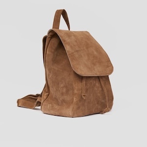Puede incluir: Una mochila de ante marr&oacute;n con cierre de cord&oacute;n y solapa superior. La mochila tiene una sola correa y un peque&ntilde;o bolsillo en la parte delantera.