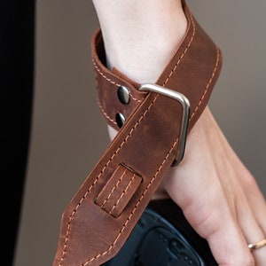 May include: A brown leather camera strap with a silver buckle and stitching. The strap is worn around a person's wrist.