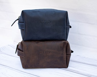 May include: Two leather toiletry bags stacked on top of each other. The top bag is navy blue and the bottom bag is brown. Both bags have the word "LIBERTY" embossed on the front.