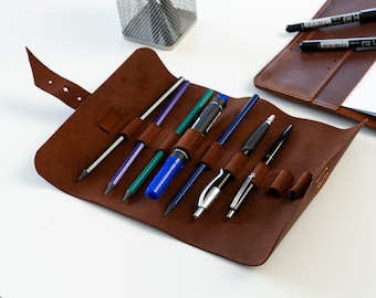 May include: Brown leather pencil case with multiple slots for pens and pencils. The case is rolled up and has a strap with a buckle. The case is filled with a variety of writing instruments, including pencils, pens, and a highlighter.