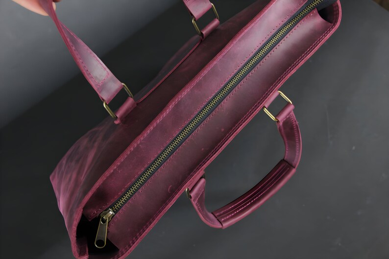 May include: A burgundy leather tote bag with a zipper closure and brass-colored hardware. The bag has two handles and a shoulder strap. The leather has a distressed finish, giving it a vintage look.