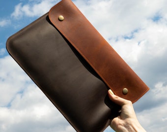 May include: A two-tone leather laptop sleeve in brown and tan. The sleeve is held by a hand against a backdrop of a cloudy blue sky. The sleeve has a flap secured by two brass-colored snaps.
