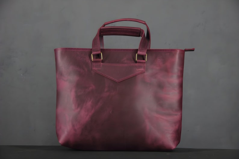 May include: A burgundy leather tote bag with a rectangular shape and two handles. The bag has a small pocket on the front and gold-colored metal hardware. The bag is made of a smooth, textured leather.
