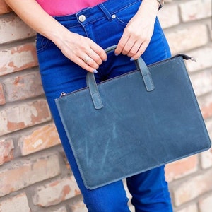 Puede incluir: Un bolso tote de cuero azul con cierre de cremallera y dos asas superiores. El bolso lo lleva una persona que lleva pantalones vaqueros azules.