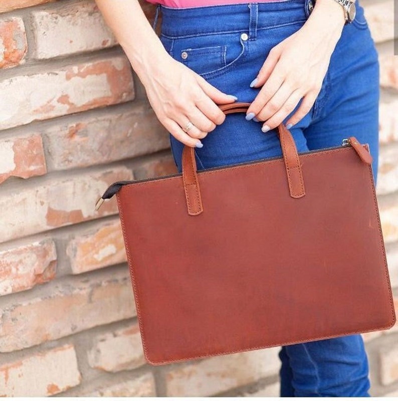 Puede incluir: Un bolso de cuero marr&oacute;n para port&aacute;til con cierre de cremallera y dos asas. El bolso lo lleva una persona que lleva pantalones vaqueros azules.