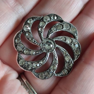 Op de afbeelding: Een zilverkleurige broche met een draaiend ontwerp van strass. De broche heeft de vorm van een bloem met meerdere lagen strass.
