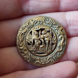 Pode incluir: Um botão de metal dourado com um design em relevo de duas figuras em um cenário medieval. O botão é circular e tem uma borda detalhada com motivos florais e de volutas.