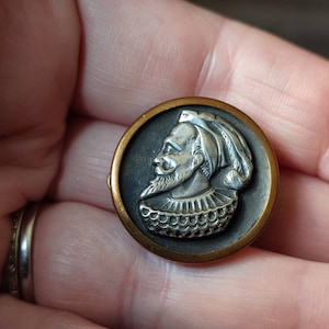 May include: A silver and gold button featuring a detailed profile of a man wearing a hat and a ruff collar.