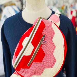 May include: A quilted fanny pack featuring a geometric pattern of red, cream, and red-and-white micro-dot fabric. The bag has a white zipper closure, a front zippered pocket with a small metallic sewing machine charm, and an adjustable strap.