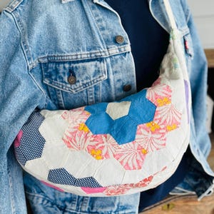 May include: A white, crescent-shaped bag with a patchwork design. The bag features hexagonal patches in blue, pink, and white with floral and polka dot patterns. It has a cream-colored strap and is worn over a denim jacket.