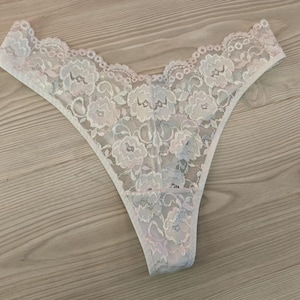 Op de afbeelding: Fijne witte kanten string met een bloemenpatroon. De randen zijn geschulpt en afgewerkt met een zachte roze tint. Het bloemmotief bevat subtiele blauwe en roze accenten, wat zorgt voor een vrouwelijke en elegante uitstraling.