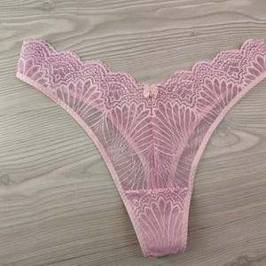 Puede incluir: Un tanga rosa claro. La delicada lencería presenta un ribete de encaje festoneado a lo largo del borde superior y detalles de encaje floral intrincados. Un pequeño lazo adorna el centro delantero. El tanga está hecho de una tela transparente y ligera.