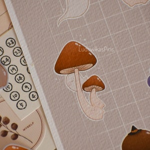 Puede incluir: Un conjunto de pegatinas de setas marrones y blancas con una pegatina de fantasma. Las pegatinas est&aacute;n sobre un fondo de cuadr&iacute;cula marr&oacute;n. El texto "LudowikasPins" est&aacute; en la esquina superior derecha de la imagen.