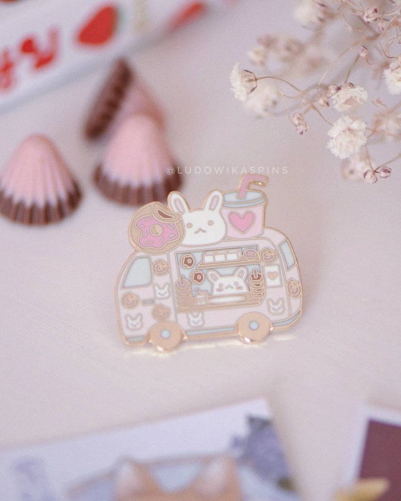 May include: A pink enamel pin featuring a cartoon food truck with a bunny on top. The truck is filled with donuts, coffee, and other treats. The pin is surrounded by white flowers.