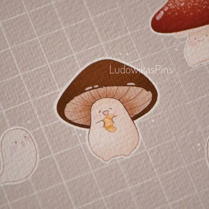 Puede incluir: Un hongo marr&oacute;n y blanco con una cara sonriente que sostiene un gusano amarillo y naranja. El hongo est&aacute; sobre un fondo de cuadr&iacute;cula marr&oacute;n claro. El texto "LudowikasPins" est&aacute; en la esquina superior derecha de la imagen.