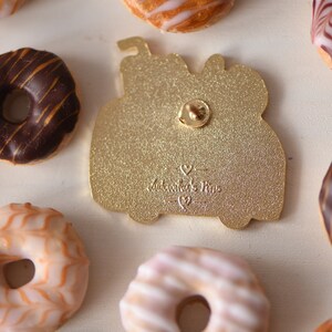 May include: Gold-colored enamel pin featuring a donut with sprinkles and a cherry on top. The pin has a glitter finish and the text "Sweetheart's Pins" on the back.