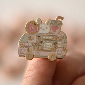 May include: A pink and gold enamel pin featuring a cartoon bunny rabbit driving a food truck. The truck is decorated with donuts, hearts, and other bunny rabbits.