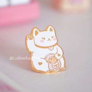 Può includere: Una spilla in smalto bianco e oro raffigurante un gatto fortunato dei cartoni animati con una zampa alzata. Il gatto ha le guance rosa e un collare a forma di cuore. La spilla include anche un gatto più piccolo e le parole "Wish You Well". La spilla è di @LudowikasPins.