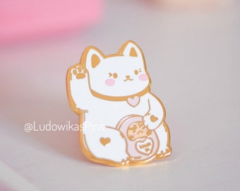 May include: A white and gold enamel pin featuring a cartoon lucky cat with a raised paw. The cat has pink cheeks and a heart-shaped collar. The pin also includes a smaller cat and the words "Wish You Well". The pin is from @LudowikasPins.