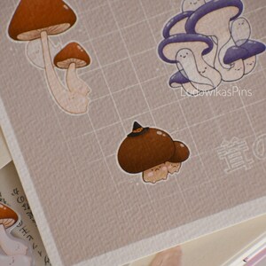Puede incluir: Un conjunto de cuatro pegatinas con forma de hongo con caras lindas. Las pegatinas son de varios tonos de marr&oacute;n y morado. Las pegatinas est&aacute;n sobre un fondo cuadriculado. El texto "LudowikasPins" es visible en la esquina inferior derecha de la imagen.
