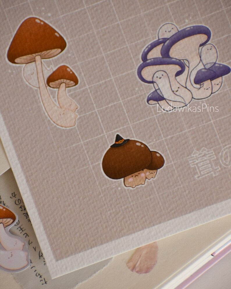 Puede incluir: Un conjunto de cuatro pegatinas con forma de hongo con caras lindas. Las pegatinas son de varios tonos de marr&oacute;n y morado. Las pegatinas est&aacute;n sobre un fondo cuadriculado. El texto "LudowikasPins" es visible en la esquina inferior derecha de la imagen.