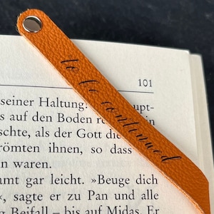 Peut inclure: Un marque-page en cuir orange textur&eacute; avec un rivet m&eacute;tallique argent&eacute; sur le dessus et le texte cursif noir to be continued imprim&eacute; en diagonale sur toute la longueur de la bande, pos&eacute; sur une page de livre ouverte avec du texte imprim&eacute;.