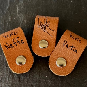 May include: Three brown leather tags with silver snaps. One tag reads "bester Neffe", another features a hand-holding illustration, and the third says "beste Patin". The tags are on a dark surface.