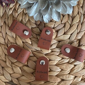 May include: Five small, brown leather tags with silver snaps. Each tag has the name "BreiNesen" and a small heart symbol printed on it. The tags are arranged on a woven, light brown surface.