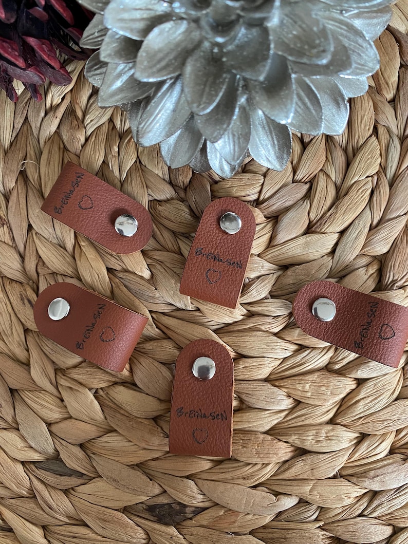 May include: Five small, brown leather tags with silver snaps. Each tag has the name "BreiNesen" and a small heart symbol printed on it. The tags are arranged on a woven, light brown surface.