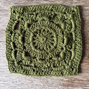 May include: A square crochet motif in a solid olive green color. The design features a circular center with radiating stitches, transitioning into a square shape with openwork details and a textured border created by various yarn loops.