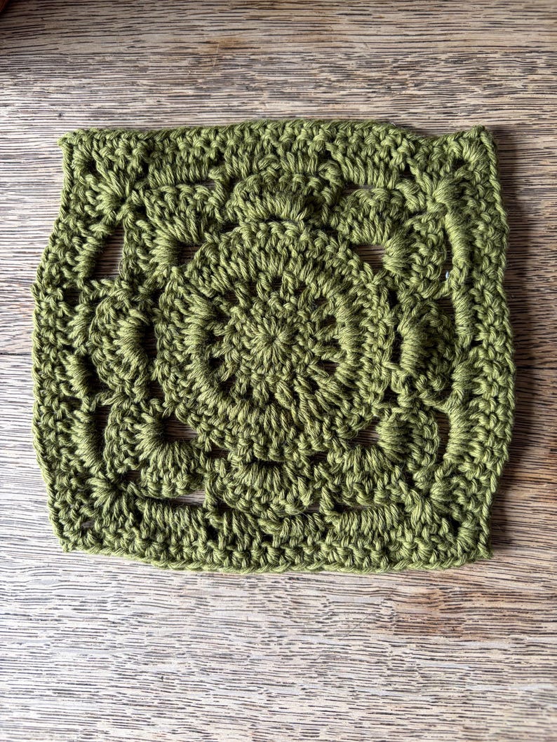 May include: A square crochet motif in a solid olive green color. The design features a circular center with radiating stitches, transitioning into a square shape with openwork details and a textured border created by various yarn loops.