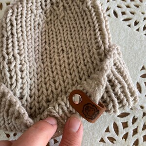 May include: A beige knitted hat with a textured pattern. The hat has a small brown leather tag with a circular design attached to the side. The hat is made of a soft, warm material.