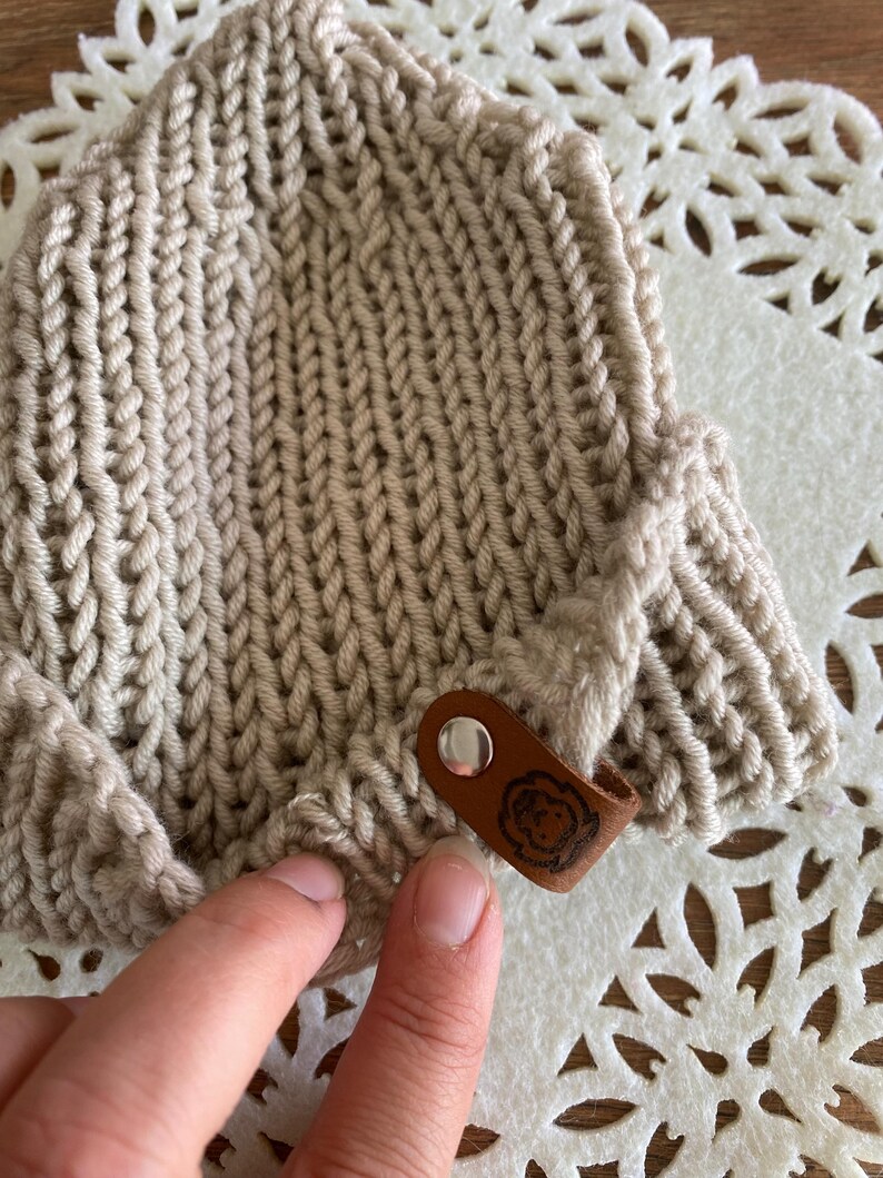 May include: A beige knitted hat with a textured pattern. The hat has a small brown leather tag with a circular design attached to the side. The hat is made of a soft, warm material.